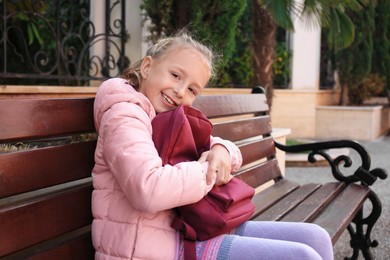Photo of Cute little girl with backpack on bench outdoors. Space for text