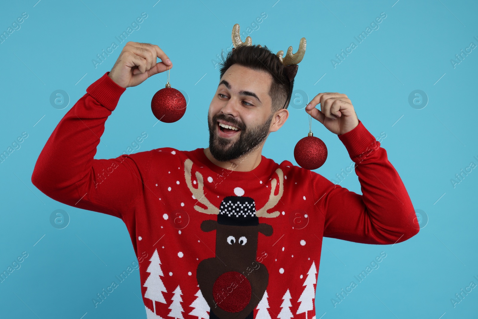 Photo of Happy young man in Christmas sweater and reindeer headband holding festive baubles on light blue background