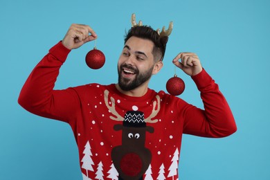 Photo of Happy young man in Christmas sweater and reindeer headband holding festive baubles on light blue background