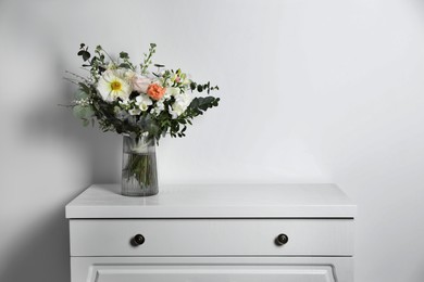 Photo of Bouquet with beautiful flowers in vase on chest of drawers near white wall. Space for text