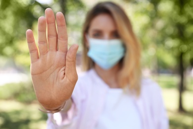 Photo of Woman in protective face mask showing stop gesture in park, focus on hand. Prevent spreading of coronavirus