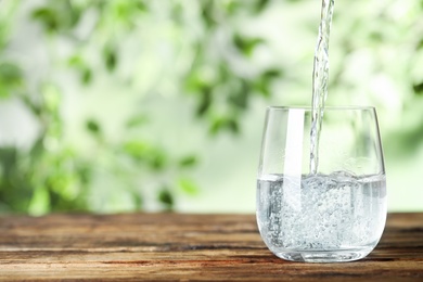 Photo of Pouring water into glass on wooden table outdoors, space for text. Refreshing drink