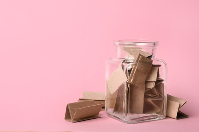 Photo of Glass jar full of brown paper sheets on pink background, space for text