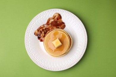 Photo of Delicious pancakes with butter, maple syrup and fried bacon on light green background, top view