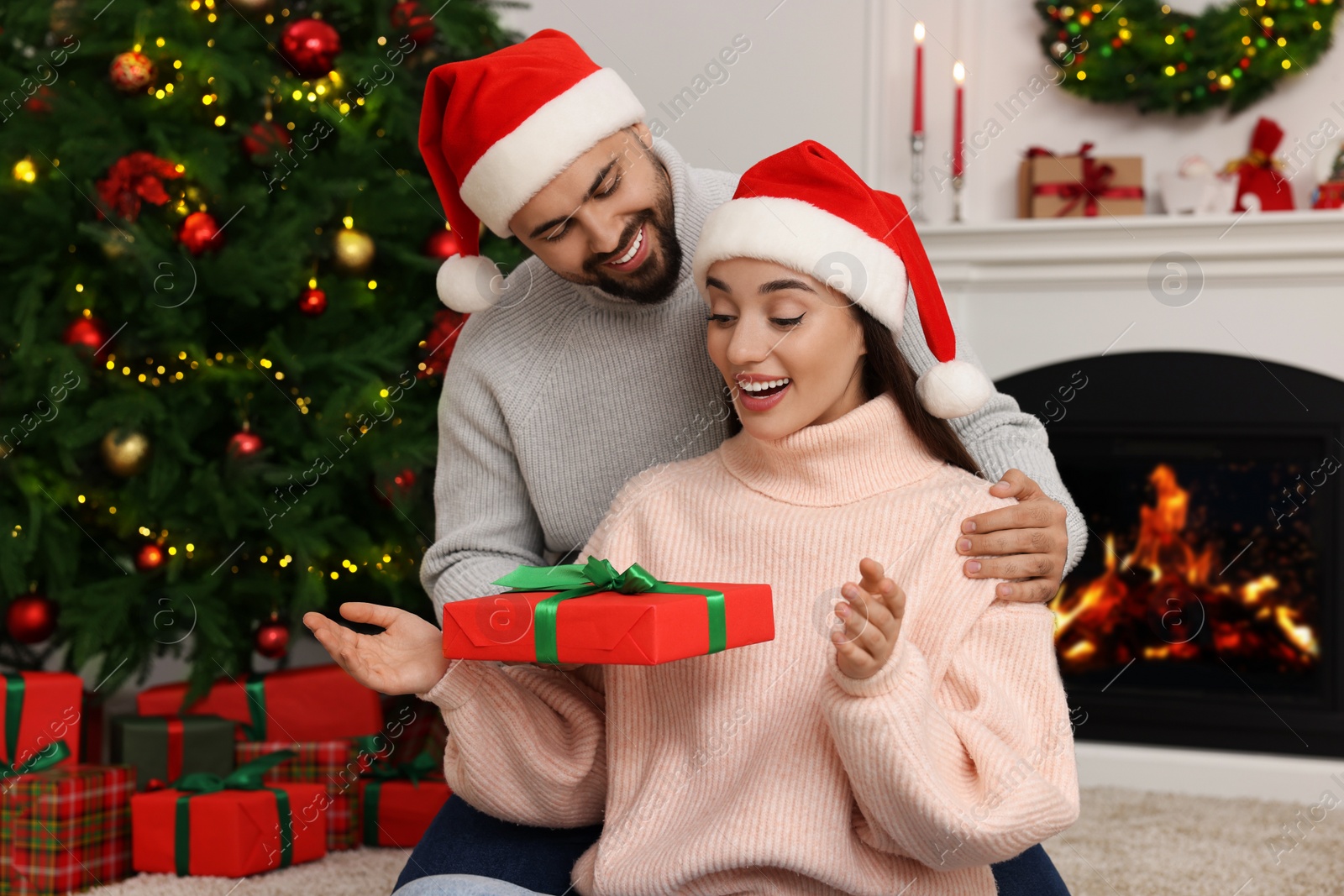Photo of Happy man in Santa hat presenting Christmas gift to his surprised girlfriend at home