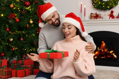 Happy man in Santa hat presenting Christmas gift to his surprised girlfriend at home