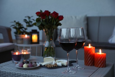 Glasses of wine, vase with roses, burning candles and snacks on rattan table at balcony in evening, closeup