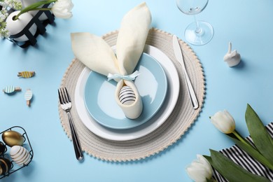 Festive table setting with painted eggs, plates and white tulips on light blue background, above view. Easter celebration