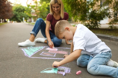 Nanny with cute little boy drawing house with chalks on asphalt