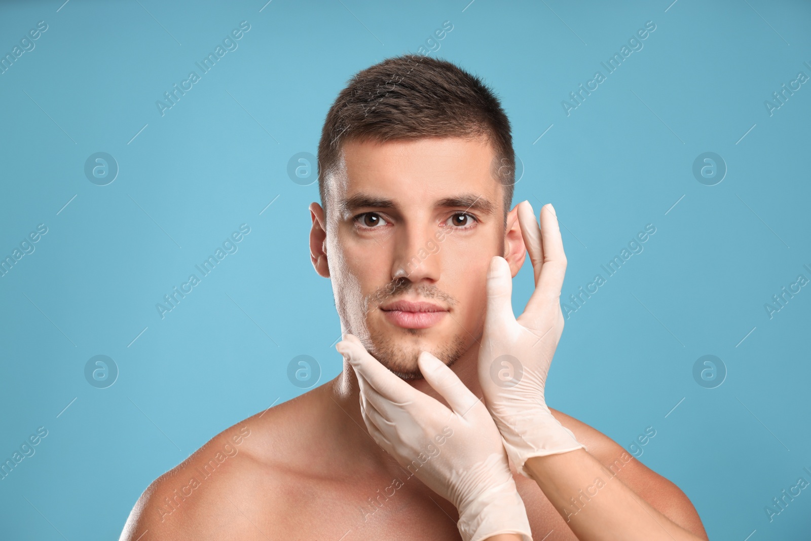 Photo of Doctor examining man's face for cosmetic surgery on light blue background