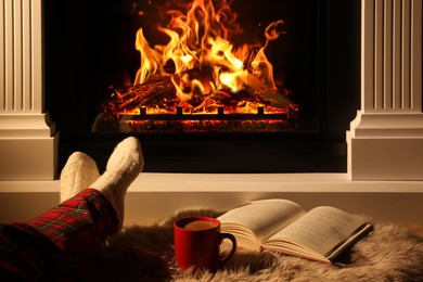 Photo of Woman with cup of drink and book near fireplace at home, closeup