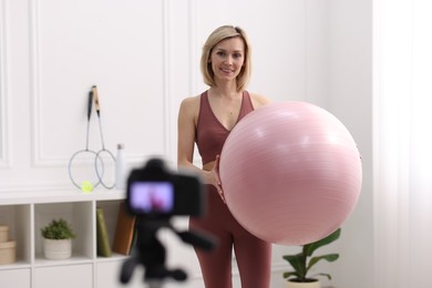 Smiling sports blogger holding fit ball while recording fitness lesson with camera at home