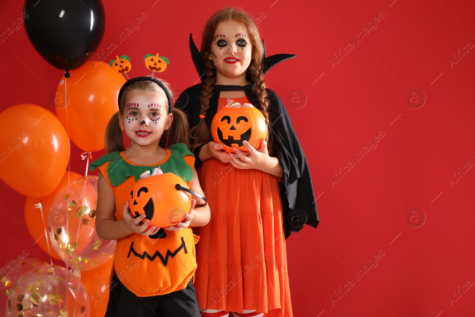 Photo of Cute little kids with pumpkin candy buckets and balloons wearing Halloween costumes on red background