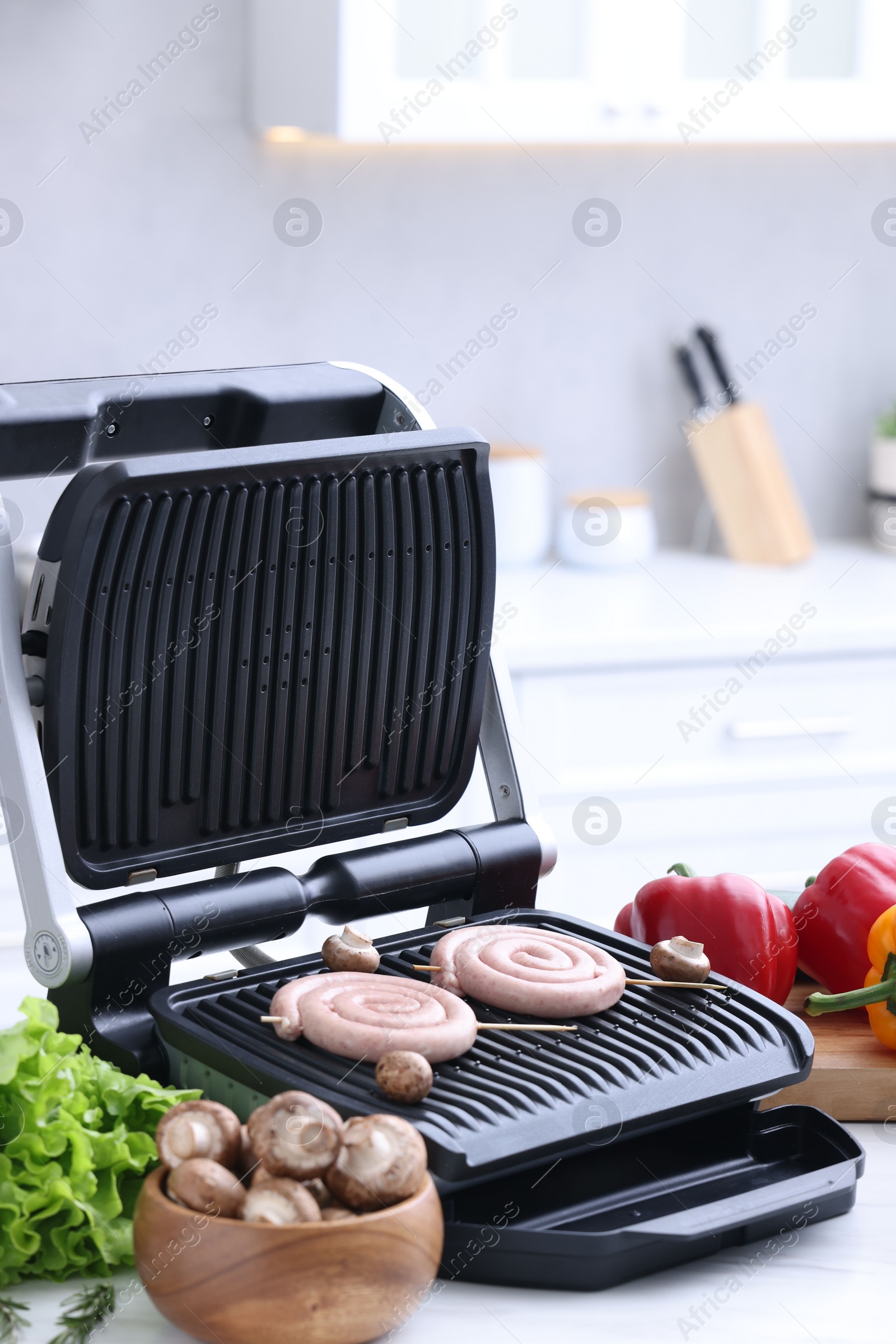 Photo of Electric grill with homemade sausages, mushrooms, and vegetables on white table in kitchen