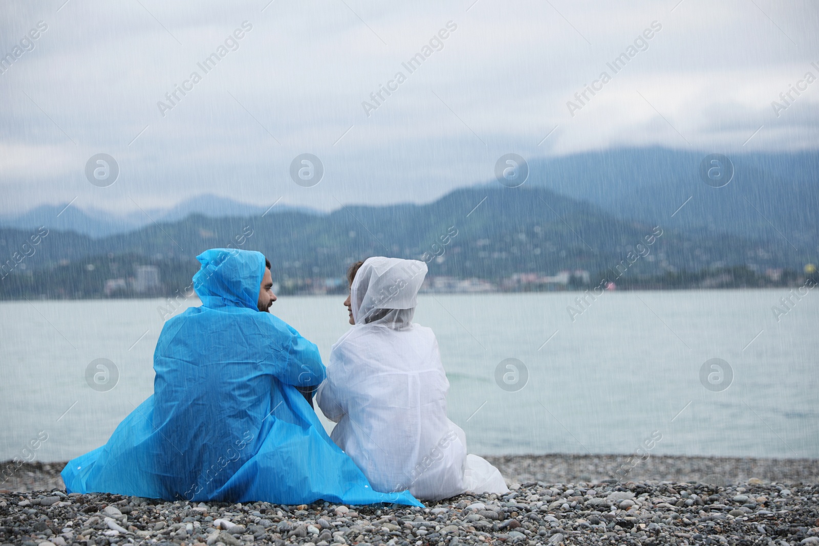 Photo of Young couple in raincoats enjoying time together under rain on beach, back view. Space for text