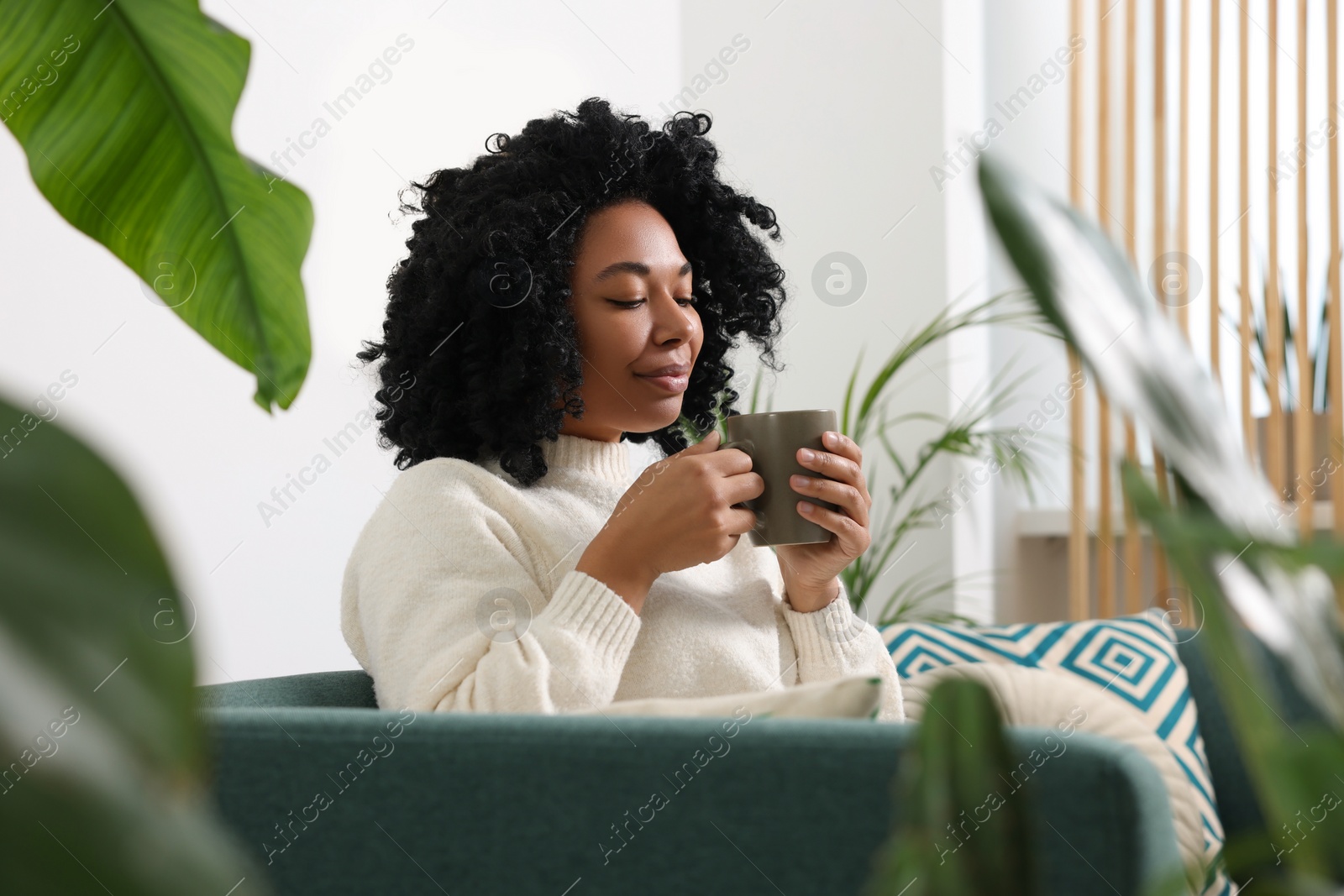 Photo of Relaxing atmosphere. Woman with cup of hot drink on sofa near beautiful houseplants in room