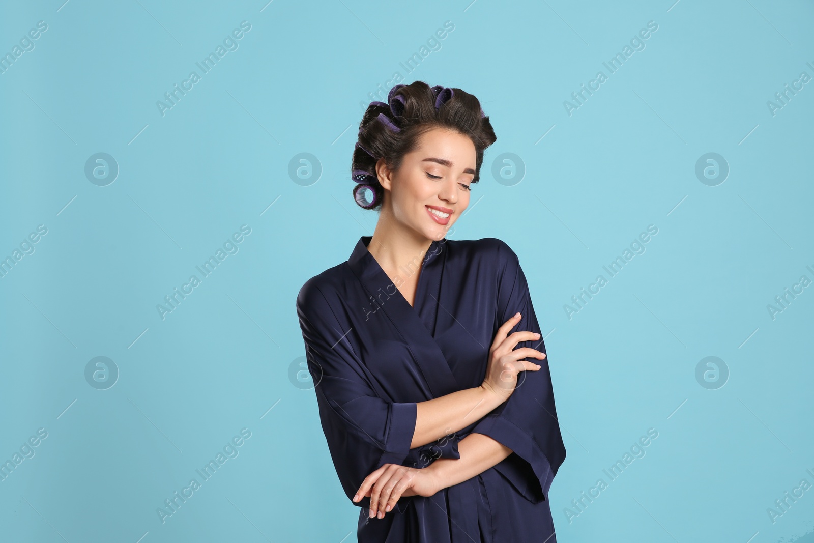 Photo of Happy young woman in silk bathrobe with hair curlers on light blue background