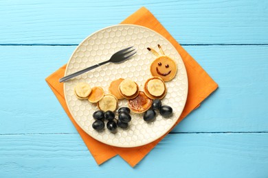 Creative serving for kids. Plate with cute caterpillar made of pancakes, grapes and banana on light blue wooden table, top view