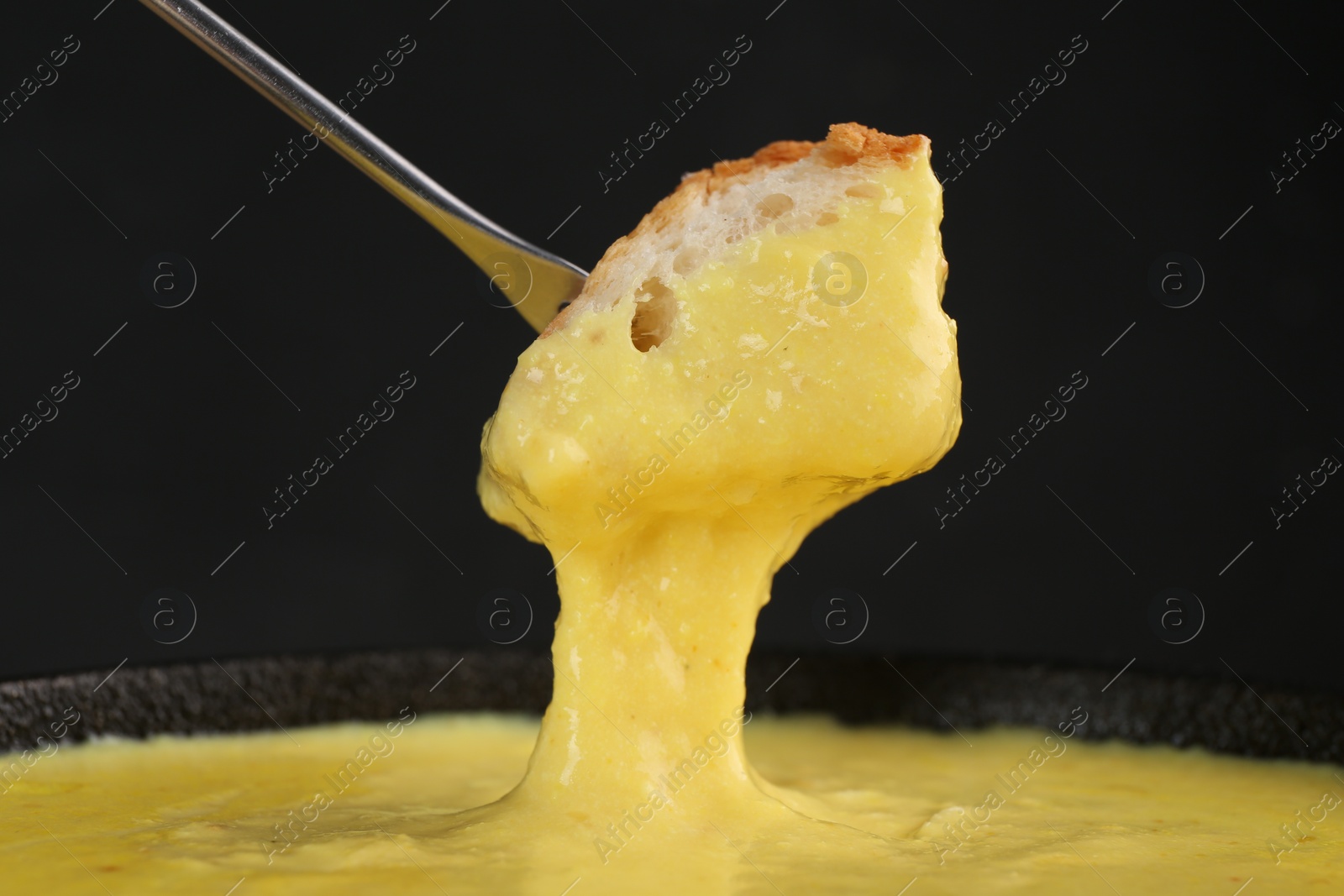 Photo of Dipping piece of bread into fondue pot with tasty melted cheese against dark gray background, closeup