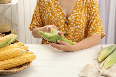 Woman husking corn cob at white wooden table, closeup