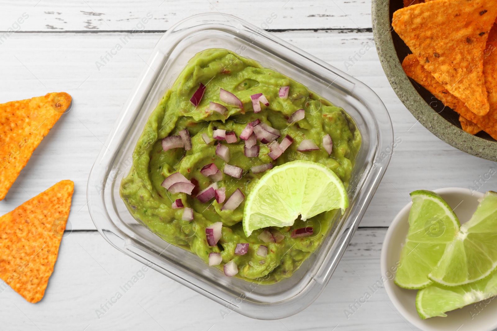 Photo of Bowl of delicious guacamole with onion, lime and nachos chips on white wooden table, flat lay