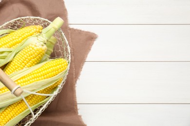 Tasty sweet corn cobs in metal basket on white wooden table, top view. Space for text