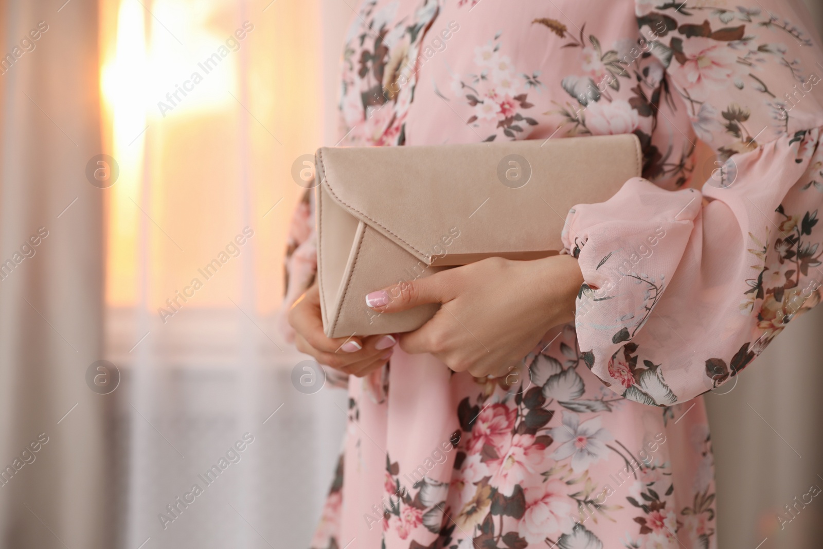 Photo of Young woman wearing floral print dress with clutch near window at home, closeup