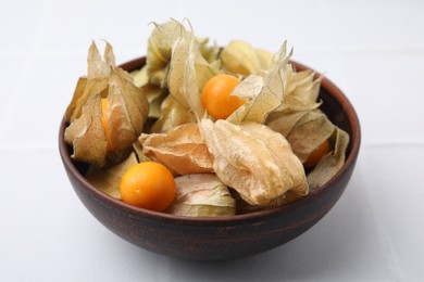 Photo of Ripe physalis fruits with calyxes in bowl on white tiled table, closeup