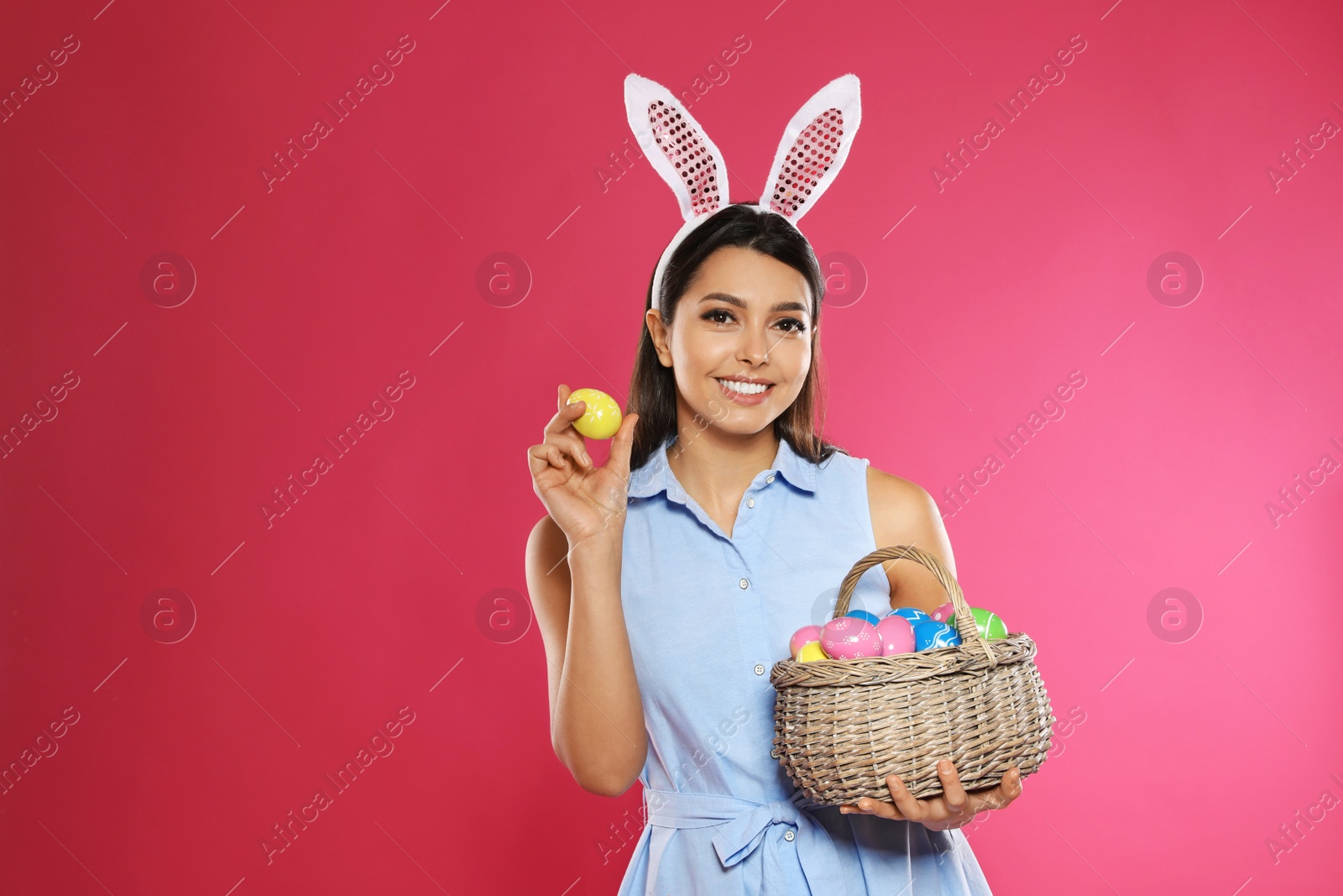 Photo of Beautiful woman in bunny ears headband holding basket with Easter eggs on color background