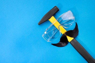 Man's face made of artificial mustache, safety glasses and hammer on blue background, top view. Space for text