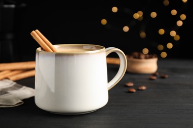 Cup of hot coffee with aromatic cinnamon on black wooden table against blurred lights. Space for text