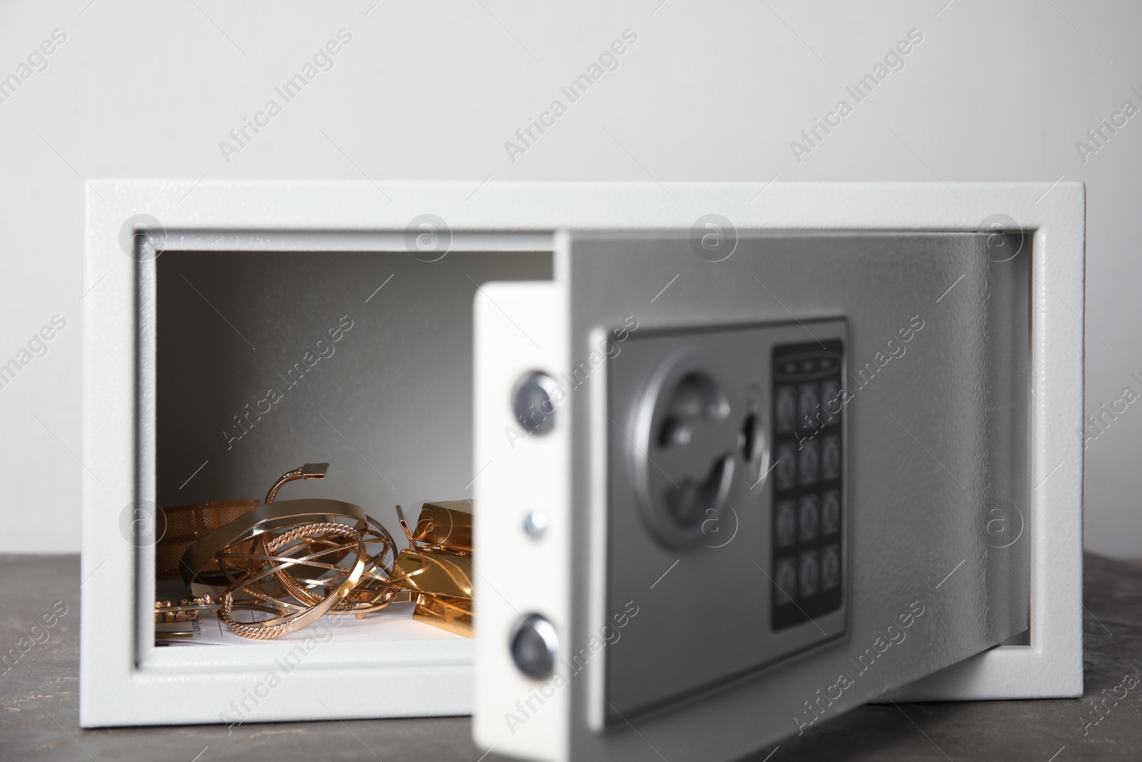 Photo of Open steel safe with gold bars and jewelry on grey table against light background