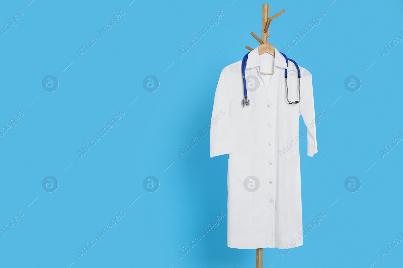 Photo of White medical uniform and stethoscope hanging on rack against light blue background. Space for text