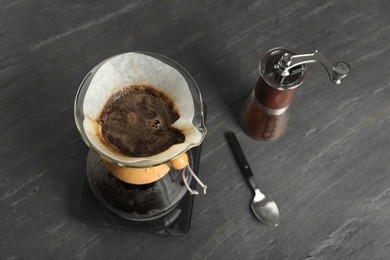 Photo of Glass chemex coffeemaker with paper filter, coffee grinder and spoon on gray table, above view