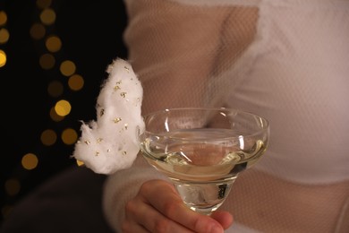 Photo of Woman holding glass of cocktail decorated with tasty cotton candy on black background with blurred lights, closeup