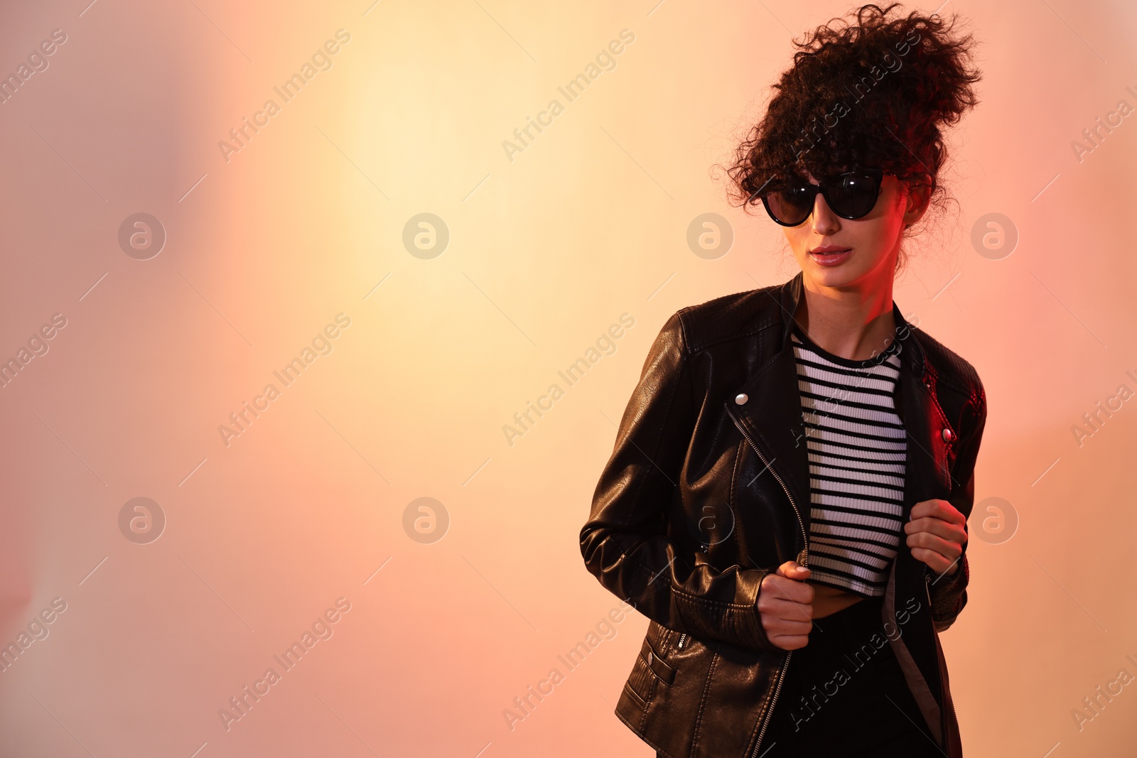 Photo of Beautiful young woman in black leather jacket and sunglasses posing on color background in neon lights. Space for text