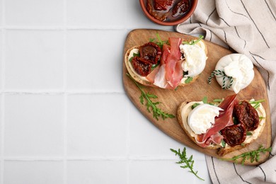 Delicious sandwiches with burrata cheese, ham and sun-dried tomatoes served on white tiled table, flat lay. Space for text