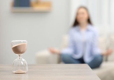 Hourglass with flowing sand on desk. Woman meditating indoors, selective focus