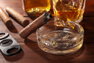 Burnt cigar, ashtray, whiskey and cutter on wooden table, closeup
