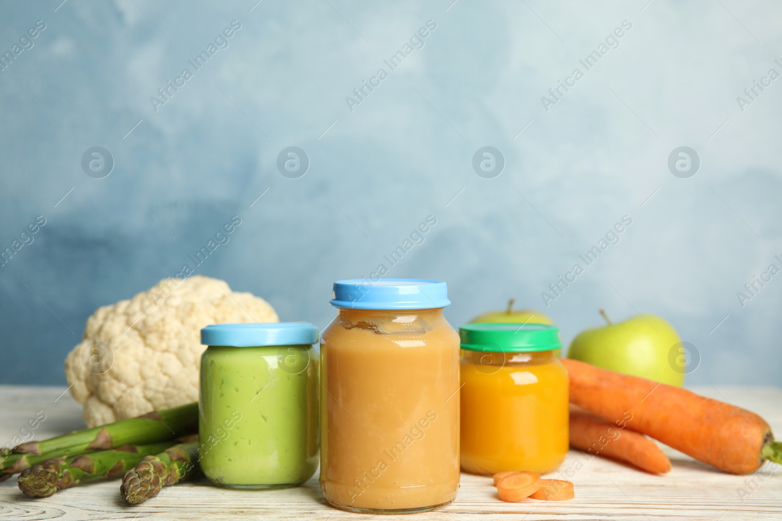 Photo of Jars with baby food and fresh ingredients on white wooden table against light blue background, space for text