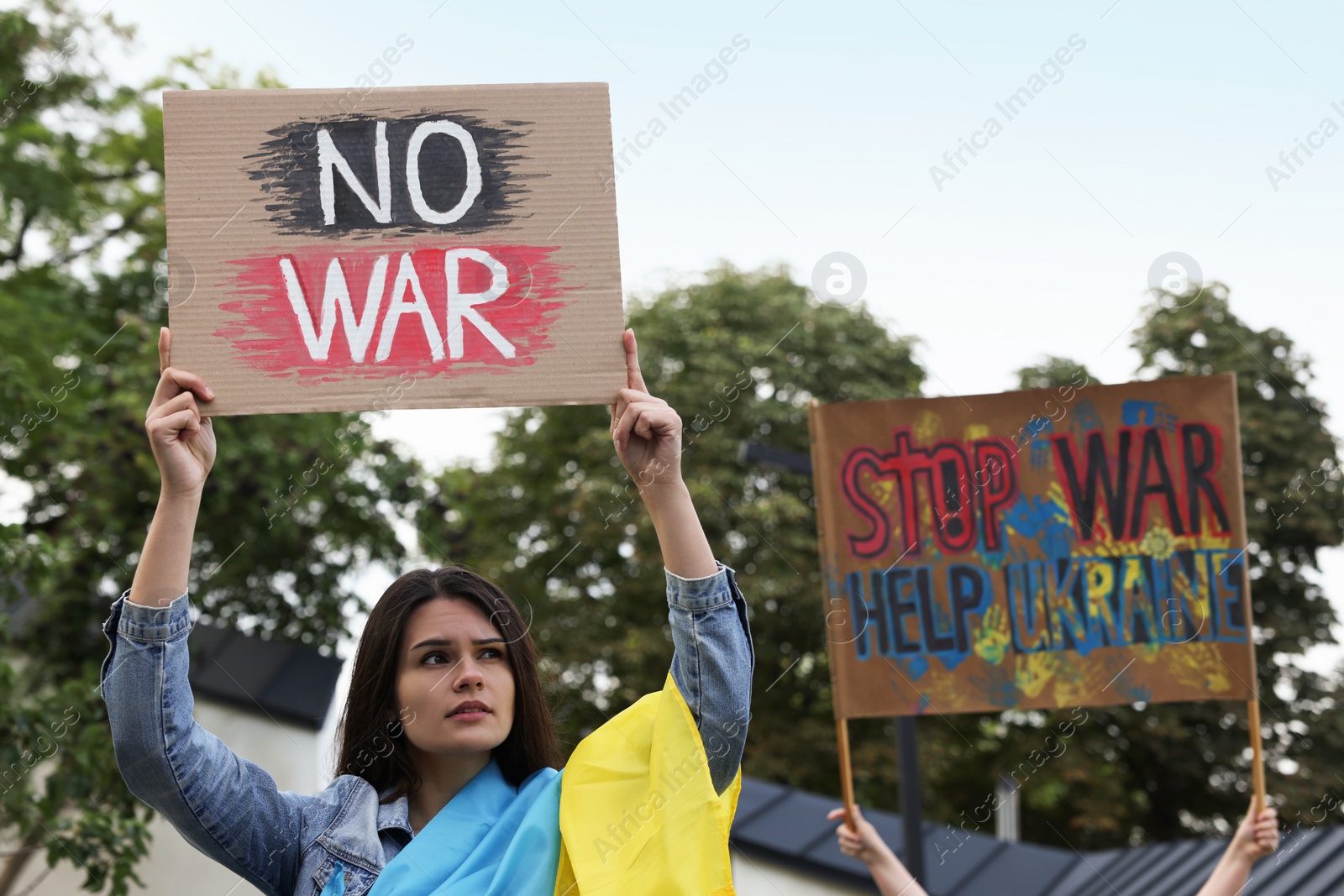 Photo of Sad woman wrapped in Ukrainian flag holding poster with words No War outdoors