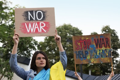 Sad woman wrapped in Ukrainian flag holding poster with words No War outdoors