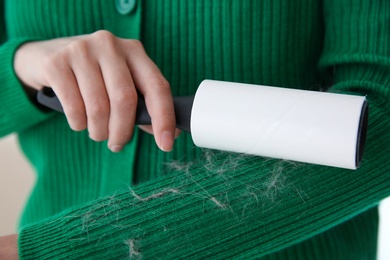 Woman removing hair from green knitted jacket with lint roller on light background, closeup