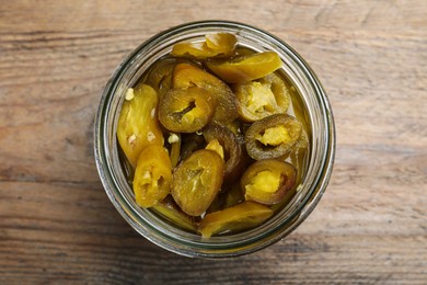 Glass jar with slices of pickled green jalapeno peppers on wooden table, top view