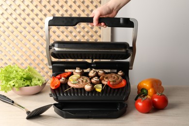 Photo of Woman cooking homemade sausages with bell peppers and mushrooms on electric grill at wooden table, closeup