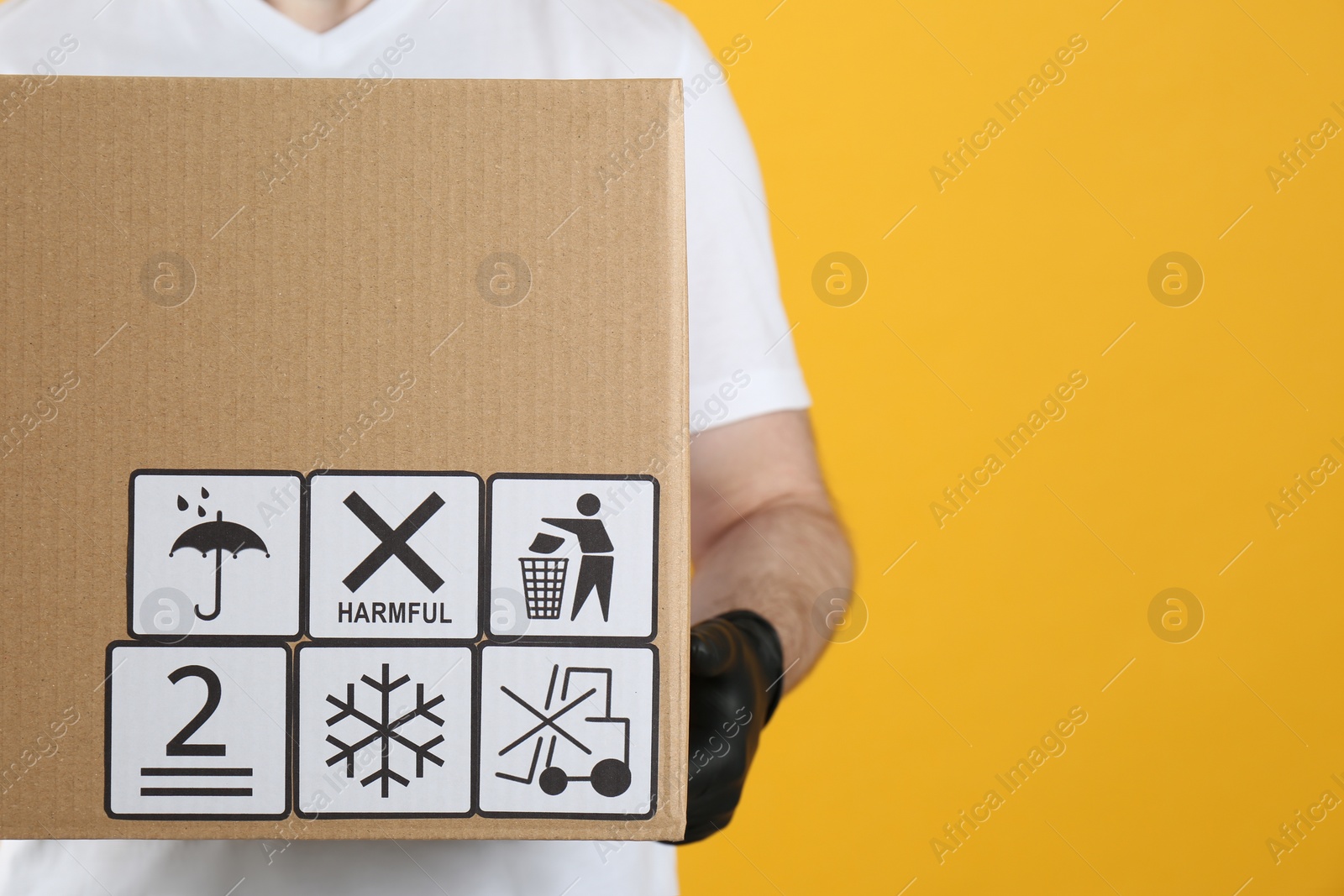 Photo of Courier holding cardboard box with different packaging symbols on yellow background, closeup. Parcel delivery