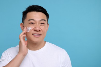 Handsome man applying cream onto his face on light blue background. Space for text