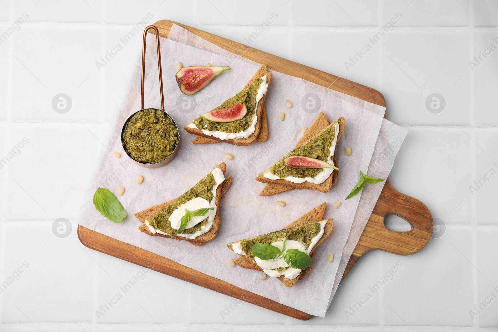 Photo of Tasty bruschettas with cream cheese, pesto sauce, figs and fresh basil on white tiled table, top view