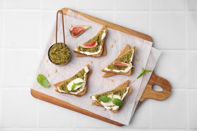 Tasty bruschettas with cream cheese, pesto sauce, figs and fresh basil on white tiled table, top view