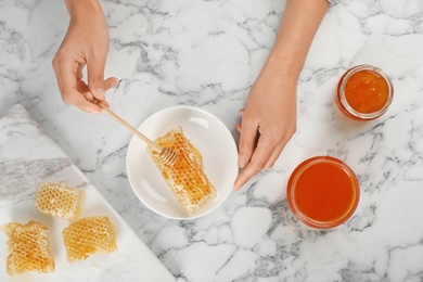 Woman pouring honey onto fresh honeycomb at marble table, top view